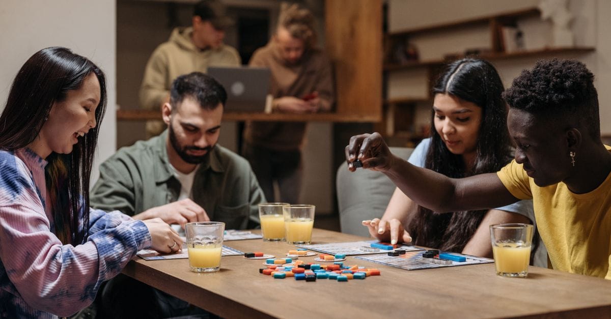 A diverse group of friends playing board games with drinks, socializing indoors.